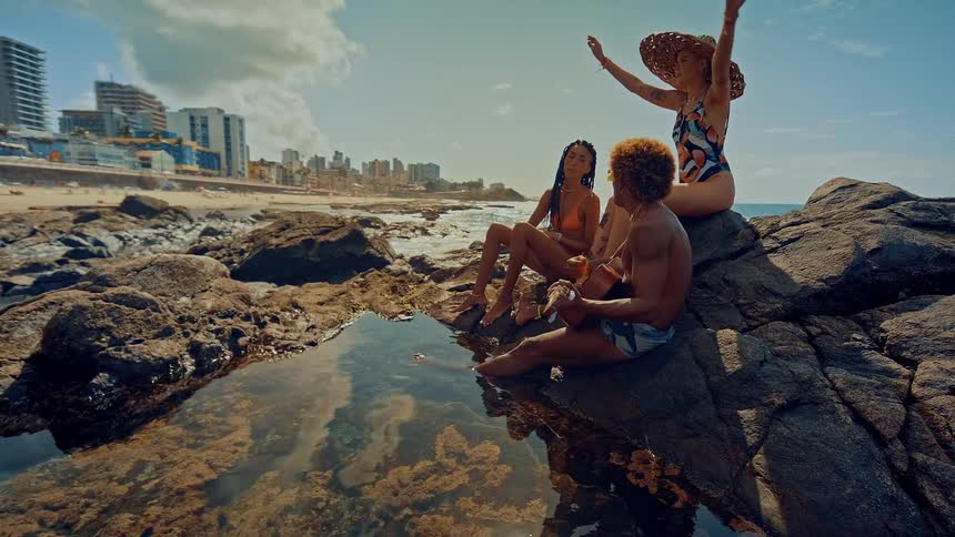 Three people in swimsuits sit on seaside rocks talking with buildings and the city visible in the background