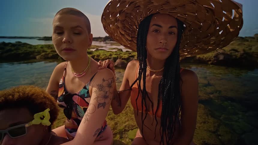 A shot from a slight low angle shows three people sitting on a rocky beach the person in the foreground has curly hair and sunglasses the person behind them has a large straw hat and tattoos and the third person has a shaved head