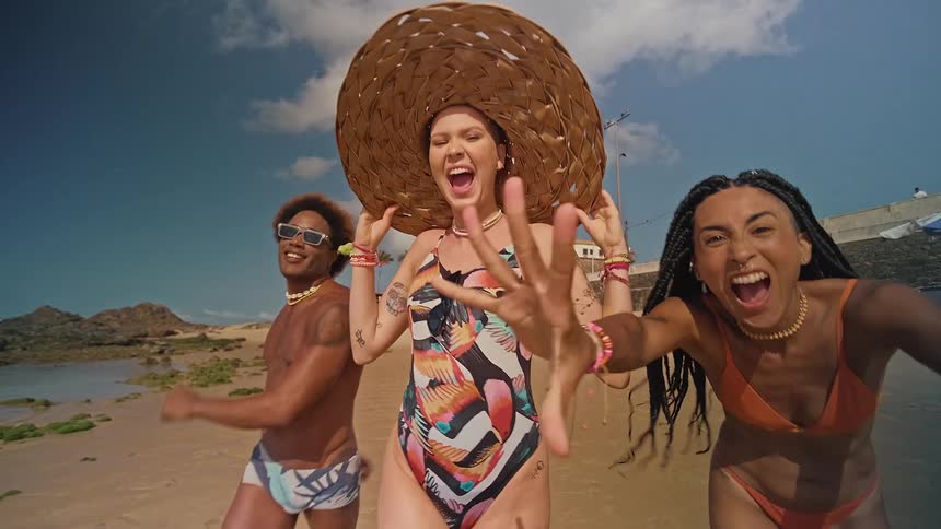 The video shows two people a man and a woman dancing and singing on a beach they are both wearing colorful clothing and smiling with a blue sky and a white building in the background