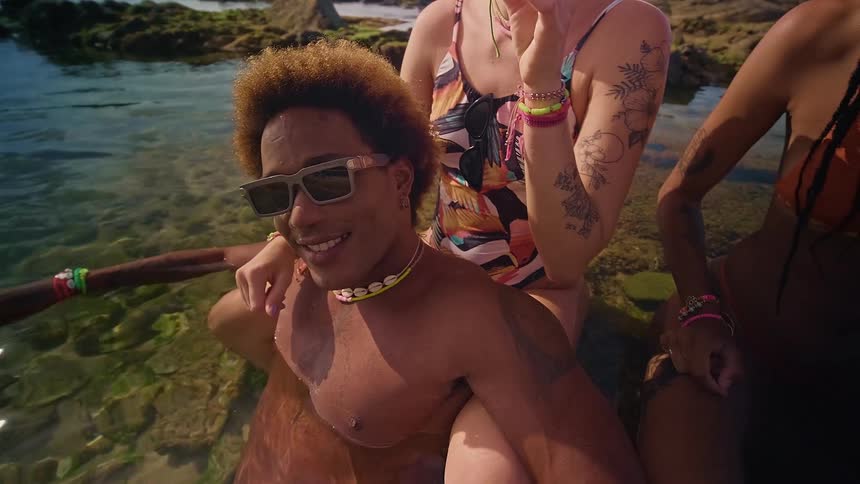 A man wearing sunglasses in the ocean water on a rocky shoreline with two women sitting behind him at midday