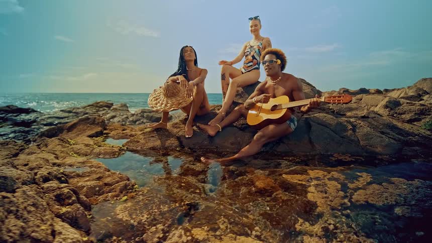 Three friends sit together on a rocky outcropping by the ocean with one of them playing a guitar under a blue sky