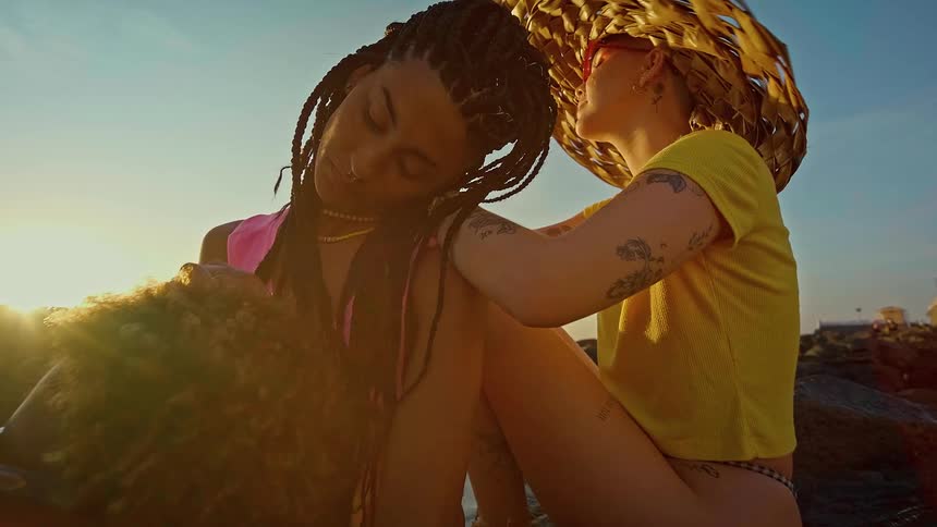 A lowangle shot on a beach shows two people sitting close together one person with braids has their eyes closed while the other person wearing a large straw hat gently places a hand on their friends cheek and looks at them