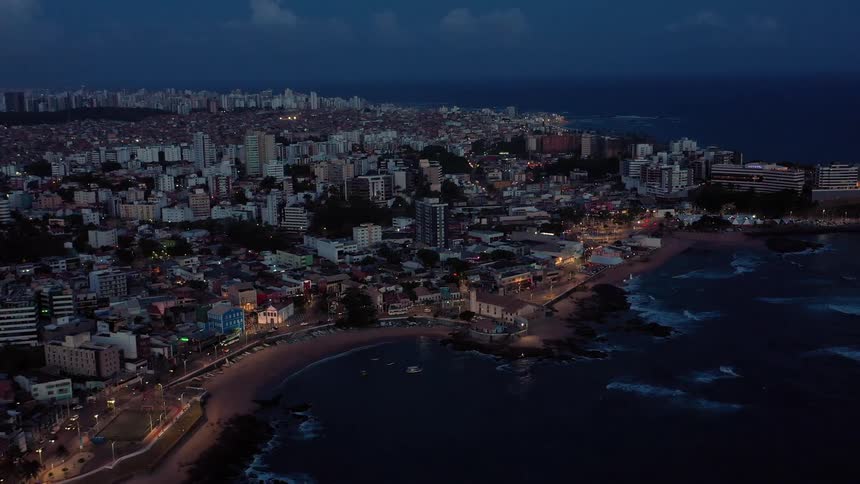 An aerial shot captures a city coastline at dusk with buildings and a street lit up along the water the sea is dark and the sky is a deep blue with the city lights creating a warm glow