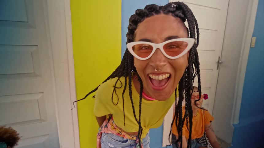 A very short video clip shows a person with braided hair and white sunglasses posing for the camera they are smiling and looking directly into the lens the background is a brightly colored wall with white and blue doors