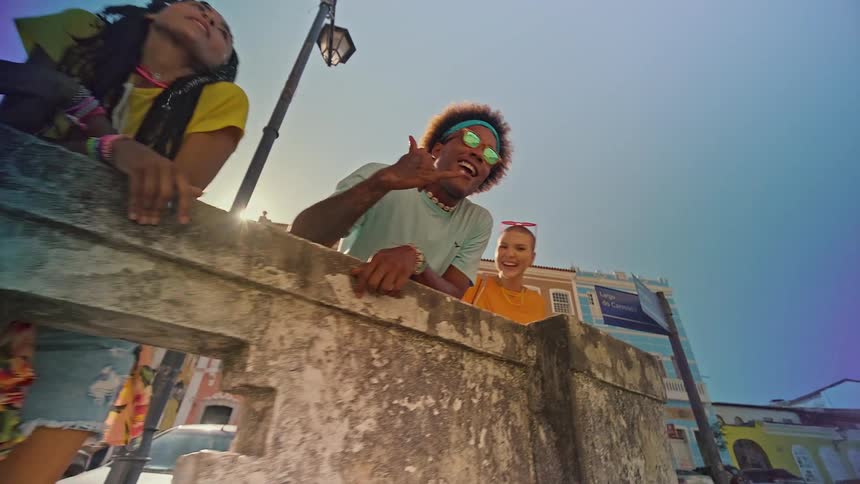 A lowangle shot shows three people leaning over a stone wall or balcony looking down the person in the middle is smiling wearing sunglasses and a headband while the others are also smiling in the bright sunshine