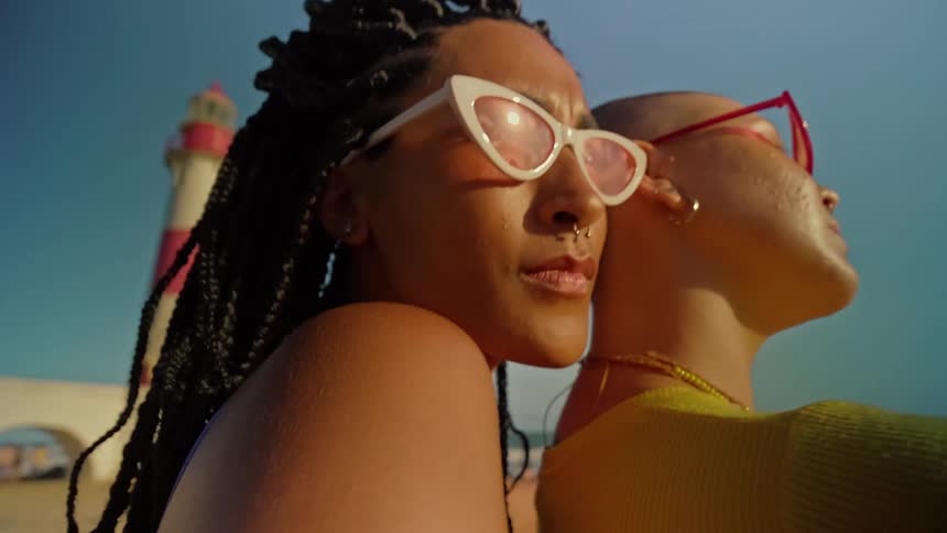 Two people stand sidebyside on a sunny beach facing away from the camera and looking out at the ocean a lighthouse is visible in the background and the person on the left has long braids while the other has a shaved head