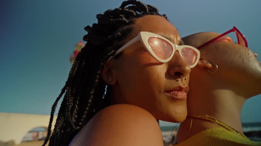 Two women wearing fashionable eyeglasses stand close together outdoors on a sunny summer day
