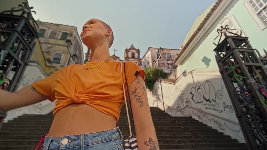 A woman with a shaved head and tattoos on her arms stands on stone stairs looking up at the sky with her hand shielding her eyes from the sun with a colorful building and a gate with ribbons visible in the background