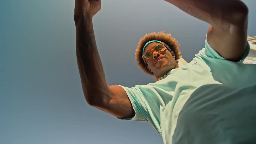 Man wearing sunglasses and a blue shirt takes a photo with his phone from a low angle on a sunny day