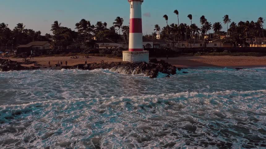 An aerial shot shows a red and white striped lighthouse on a rocky coastline next to a beach the waves are crashing on the shore and people are visible walking along the sandy beach