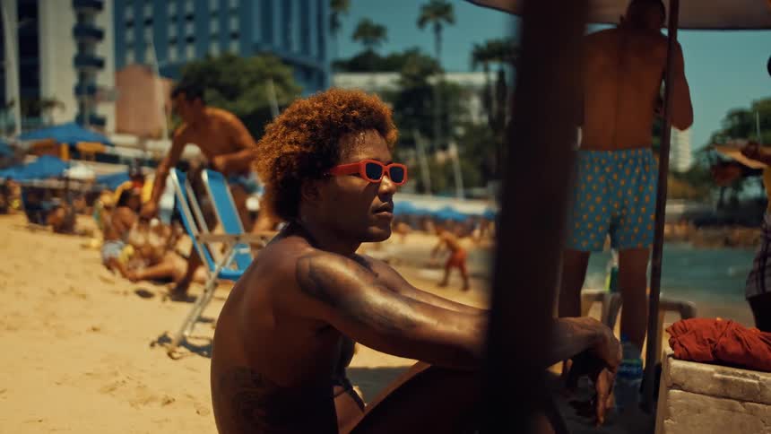 A man with curly hair and tattoos relaxes under a beach umbrella wearing bright red sunglasses surrounded by sunbathers and swimmers along a lively urban shoreline