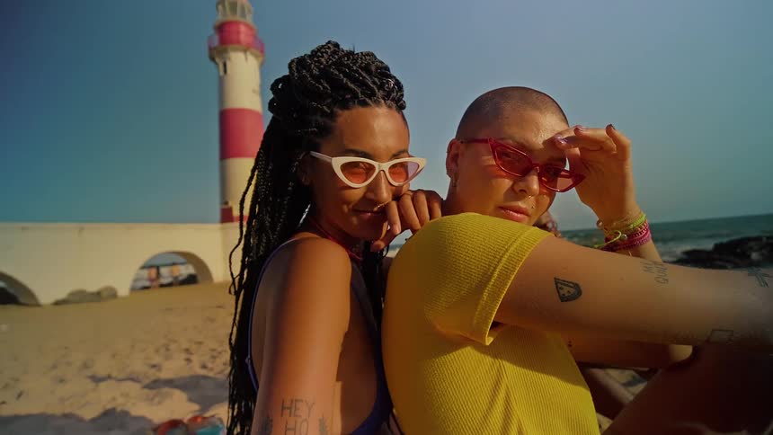 The faces of two women who are sitting on the sand of a beach and wearing sunglasses with reddish lenses and one is behind the other