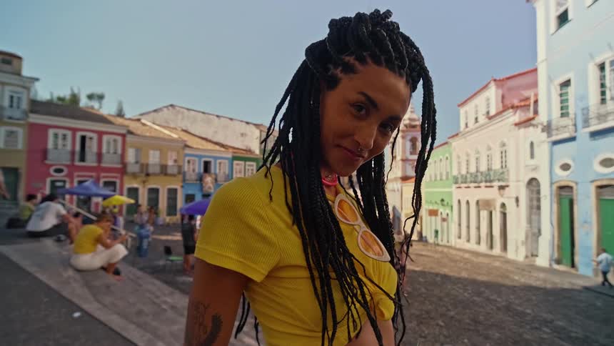 A vibrant shot of a smiling woman with dreadlocks wearing a yellow crop top and sunglasses posing confidently on a cobblestone street with a backdrop of colorful historic buildings and other people