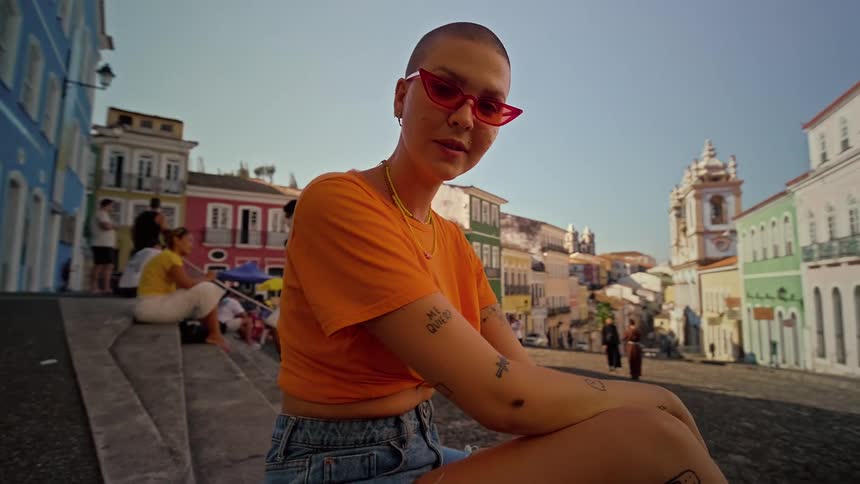 Woman wearing sunglasses and an orange tshirt sitting in a square with classic and colorful architecture
