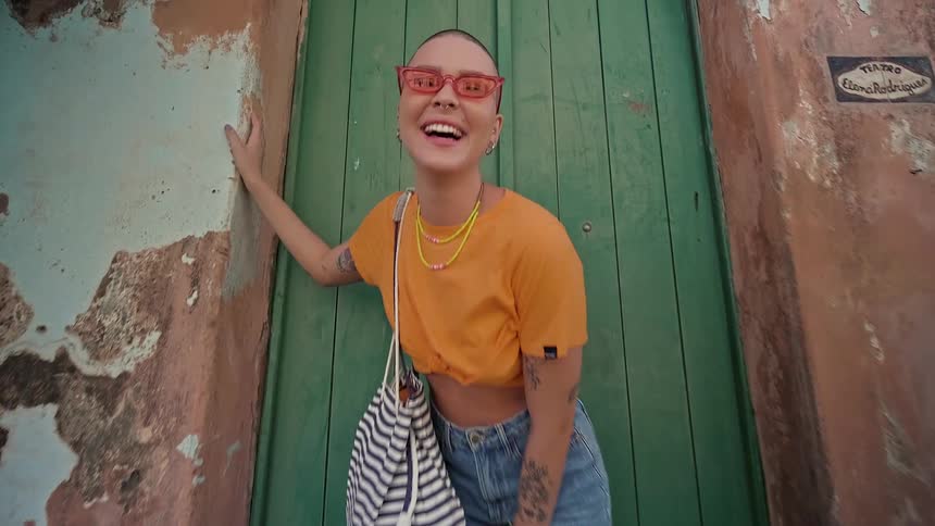 A woman with short hair and sunglasses is smiling and leaning against a door she is wearing an orange tshirt denim shorts and a striped bag over her shoulder