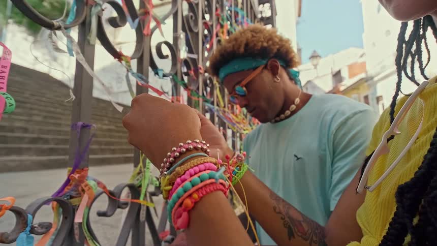 The video shows two people a man and a woman standing next to a gate covered in colorful ribbons and bracelets the man is wearing a light blue shirt a blue bandana and a necklace of seashells while the woman is wearing a yellow top with a braided ha