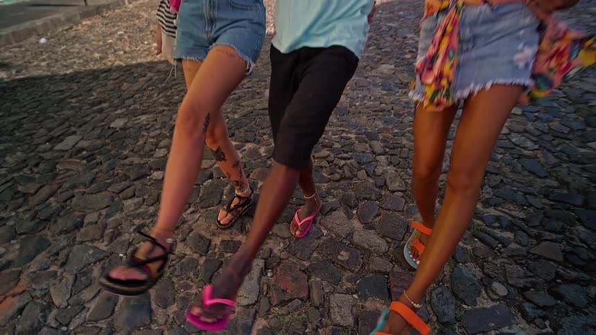 Three young people one male two female arelocked arm in arm while kicking up there feet and dancing they are smiling and are happy while they dance there way down a cobblestone street surrounded by old style colorful buildings