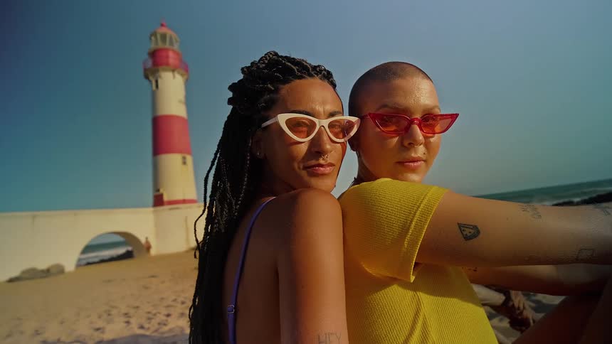 Two women sitting close together on the sand at the beach near a lighthouse