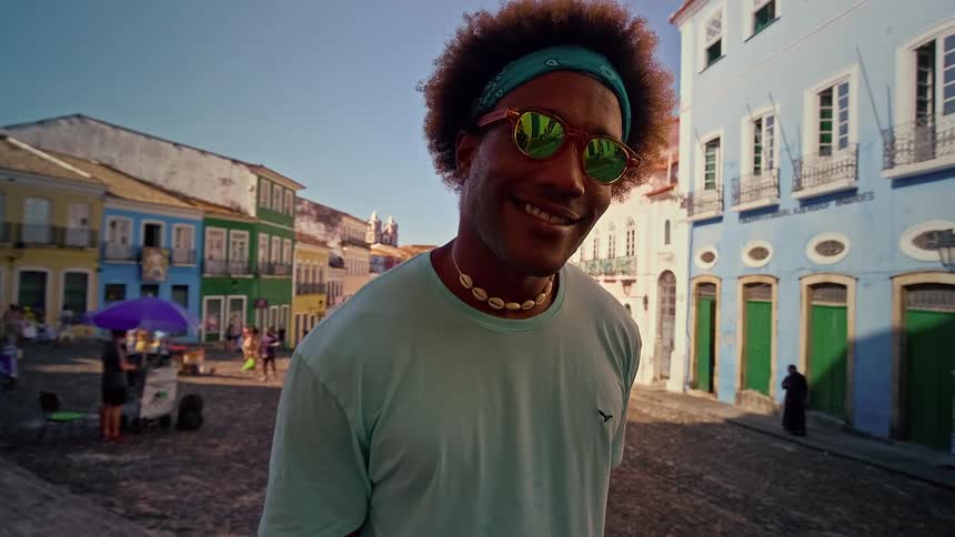 A young man standing on the sidewalk in the street wearing sunglasses with a smile on his face at midday