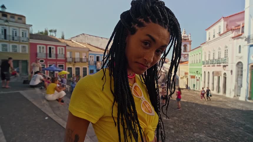 A young woman with braided hair wearing a yellow tshirt stands on the sidewalk with a smile on her face at midday
