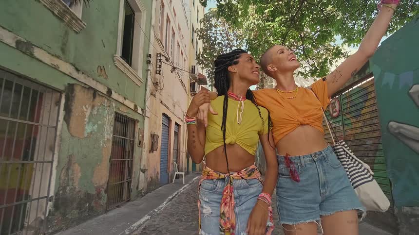 A video clip shows two young women walking down a narrow street the woman on the left has long braided hair and the woman on the right has a shaved head both are wearing cropped tshirts and denim shorts and are smiling and laughing as they walk with t