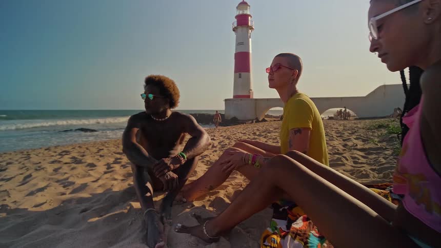 Three friends are sitting together on a sunny beach relaxing and enjoying the view of the ocean and a distant lighthouse