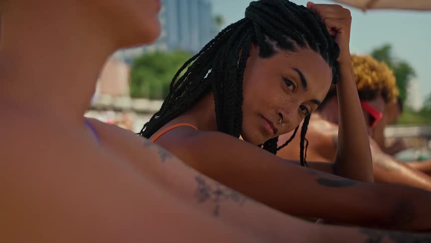 A young woman sitting beside a woman and a man on the beach at midday with a tattoo on her arm and braided hair