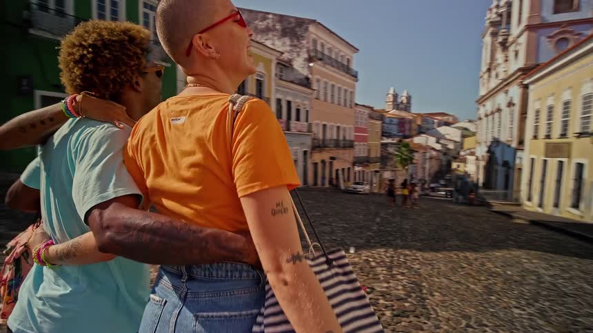 Three people are walking arminarm down a cobblestone street with a focus on their backs the street is lined with brightly colored buildings and a large church is visible in the distance
