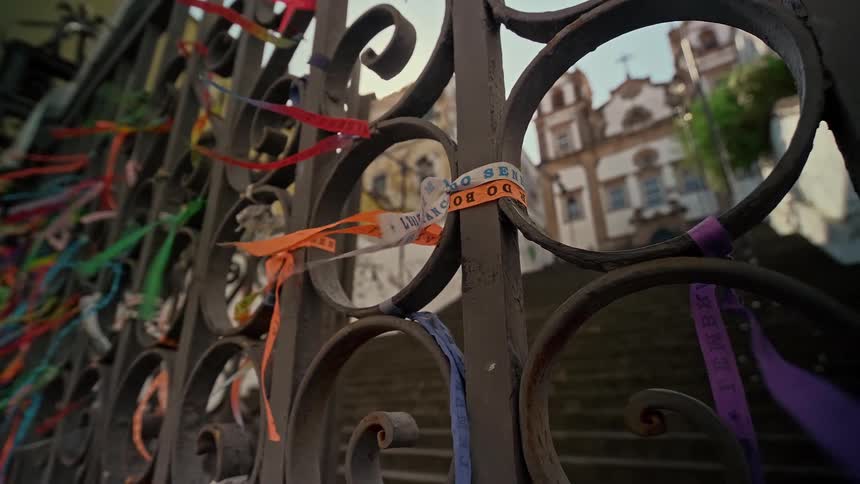 A black metal gate is covered in numerous colorful ribbons that flutter in the wind with an old church visible in the blurry background under a light blue sky