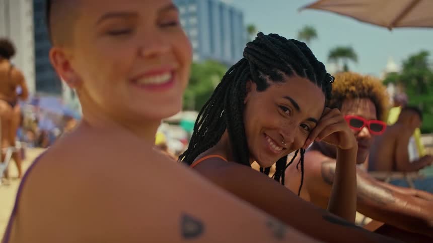 A lowangle medium shot shows three friends relaxing on a beach smiling and looking toward the camera the woman in the center with long black braids is the primary focus while the other two are slightly out of focus in the foreground and background