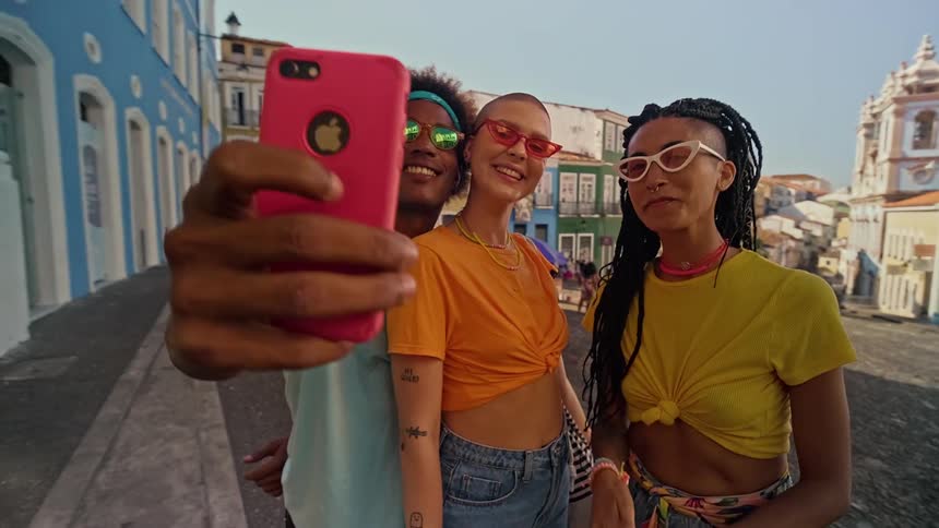 The video shows three friends taking a selfie on a cobblestone street the person on the left holds a pink phone and the group smiles and poses for the picture
