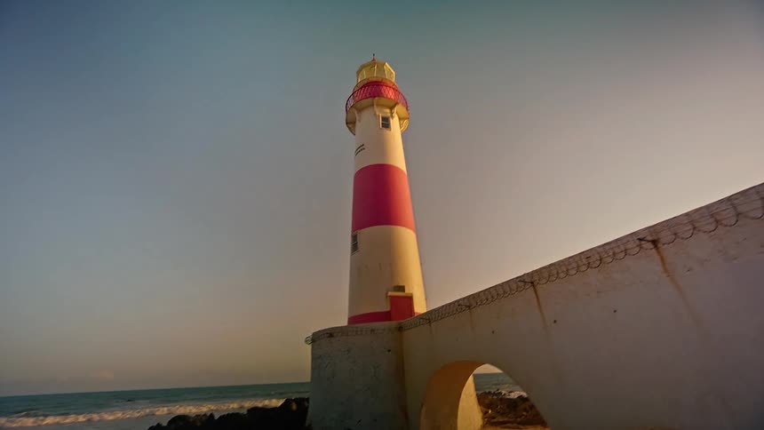 A lighthouse on the coast beside a concrete fence with ocean waves hitting the shore at midday