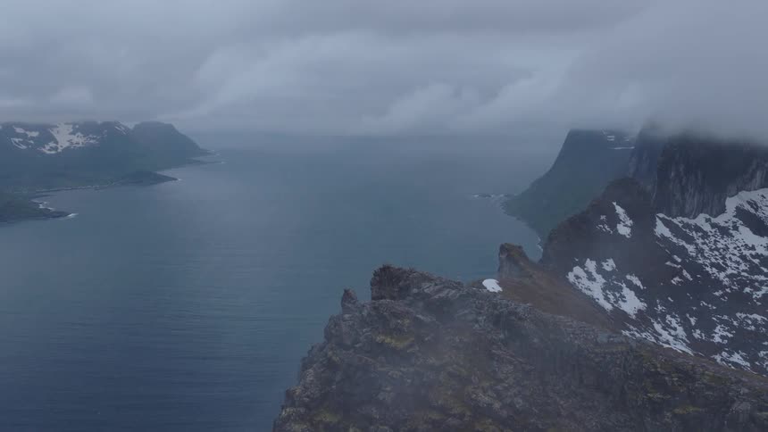 A jagged snow covered mountain ridge overlooking a fjord winding through the distant mountains under an overcast sky