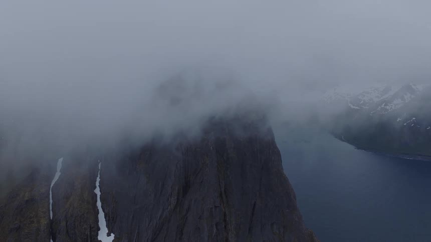 Thick fog slowly rolls in and completely obscures a tall jagged mountain peak that rises from the edge of a fjord with snow capped mountains in the distance