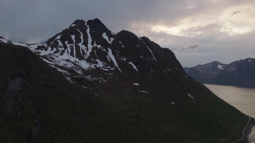 The video shows an aerial shot of a mountainous landscape with snowcapped peaks and a large body of water in the foreground the sky is dramatic with a mix of dark and light clouds
