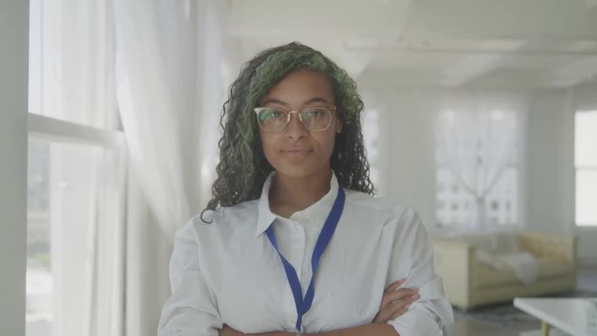 The video shows a young woman with a lanyard around her neck standing with her arms crossed she has dark hair with green highlights and is wearing a white collared shirt and glasses