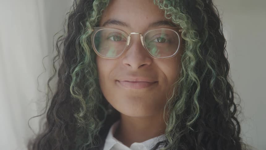 A young woman standing beside a white curtain wearing glasses with curly green and black hair smiling
