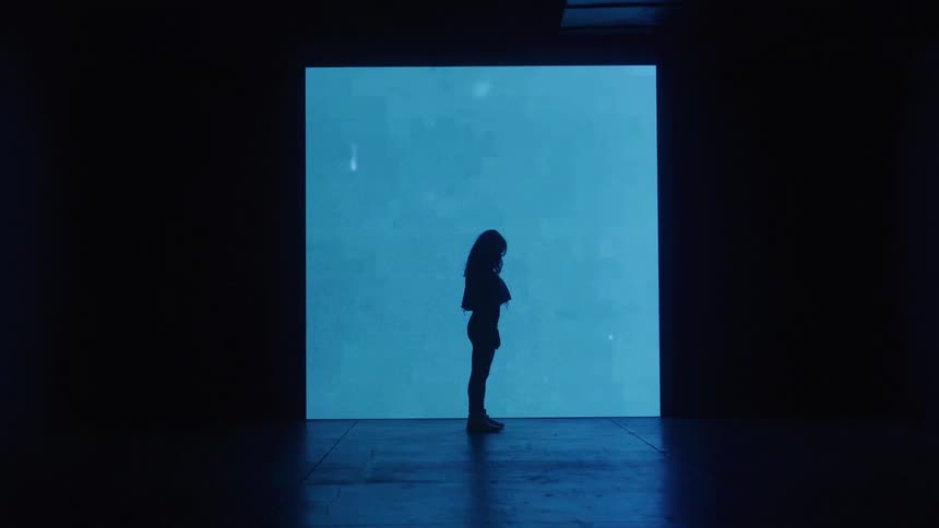 The video shows a silhouette of a person standing in front of a large screen the screens color changes from a dim light to a vibrant green then a deep red and finally back to a dim blue light