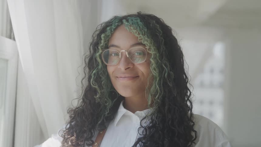 The video shows a closeup of a person with long curly dark hair with streaks of green they are wearing glasses and a white shirt and smiling at the camera