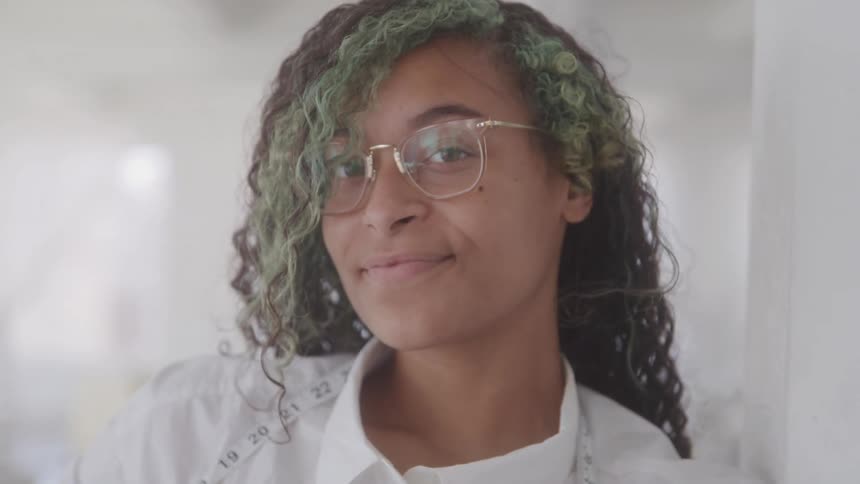A curlyhaired teenager with green and black hair a slight smile wearing square glasses and a measuring tape around her neck