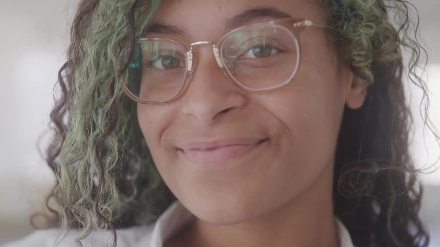 A young woman with green curly hair wearing clear glasses and smiling