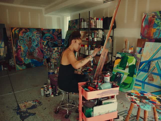 A young female artist paints a canvas while sitting in an art studio during daytime