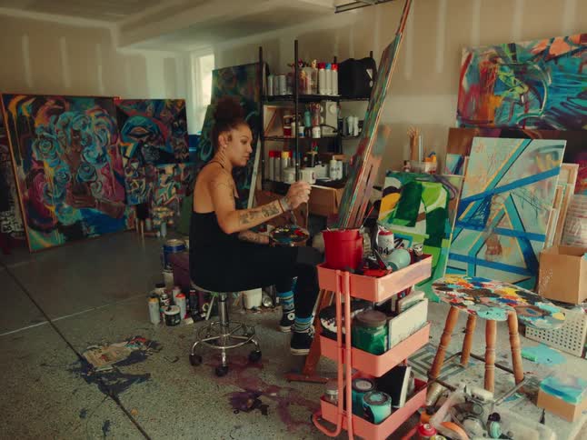 A woman with a bun and tattoos is sitting in a studio painting on a large canvas there are many colorful paintings and art supplies around her with paint splattered on the concrete floor