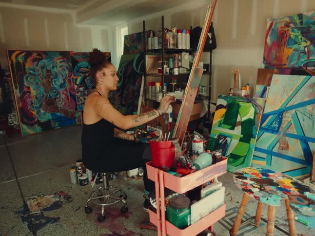 An artist wearing a black tank top and dark leggings sits on a stool and paints a large colorful canvas in a cluttered studio filled with other paintings and art supplies