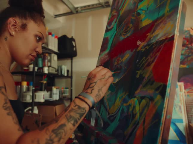 An artist with tattoos on her arms and shoulder and dark hair in a bun uses a fine paintbrush to add details to a colorful canvas in a messy studio
