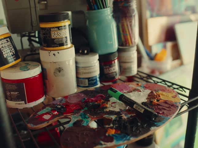 Paint palette covered in colorful paints with a marker on top surrounded by jars of art supplies on a shelf