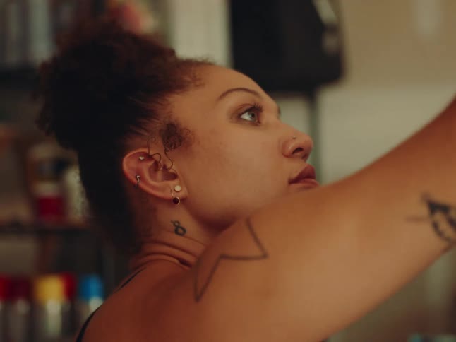 The video shows a medium closeup of a person with brown curly hair tied up in a bun the person has multiple piercings on their ears and nose