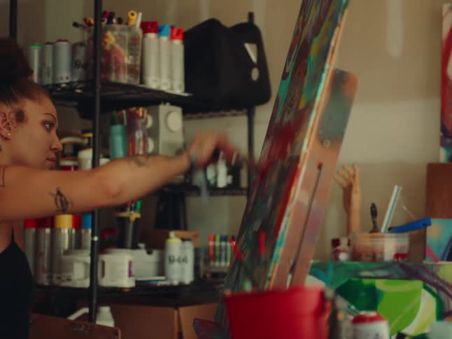 An artist with tattoos on her arms and shoulder wearing a black tank top looks at her work with a focused expression as she uses a paintbrush to add paint to a large canvas in a messy studio