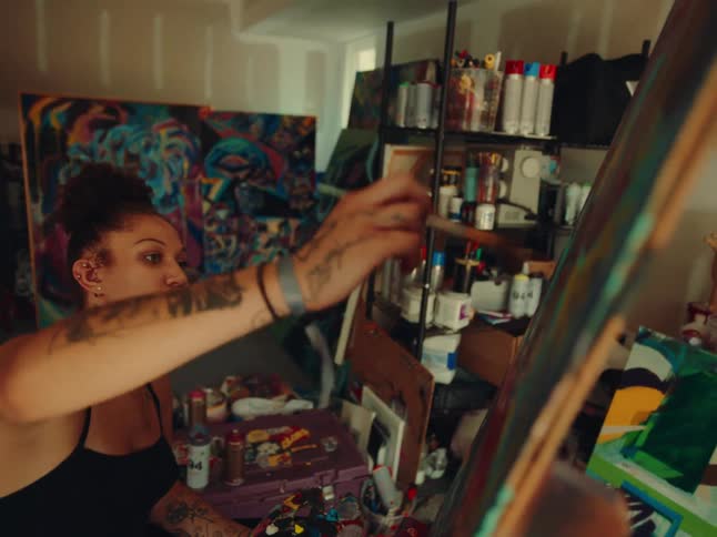 A young woman painting on a canvas inside a studio with a tattoo on her arm as the room is filled with other paintings