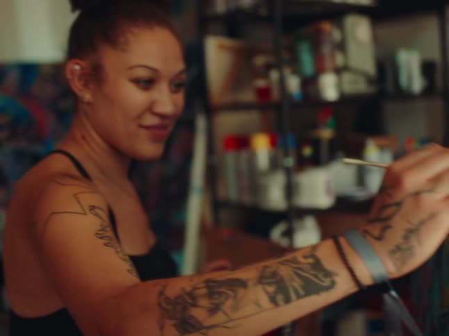 A female artist with a tattoo on her arm painting on a canvas inside a studio at midday